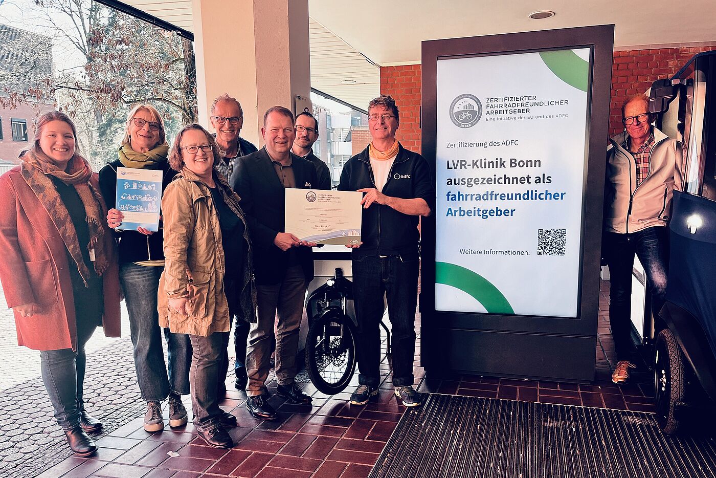 Mehrere Menschen stehen links und rechts verteilt um einen vertikalen Screen, auf dem steht "LVR-Klinik Bonn ausgezeichnet als fahrradfreundlicher Arbeitgeber".