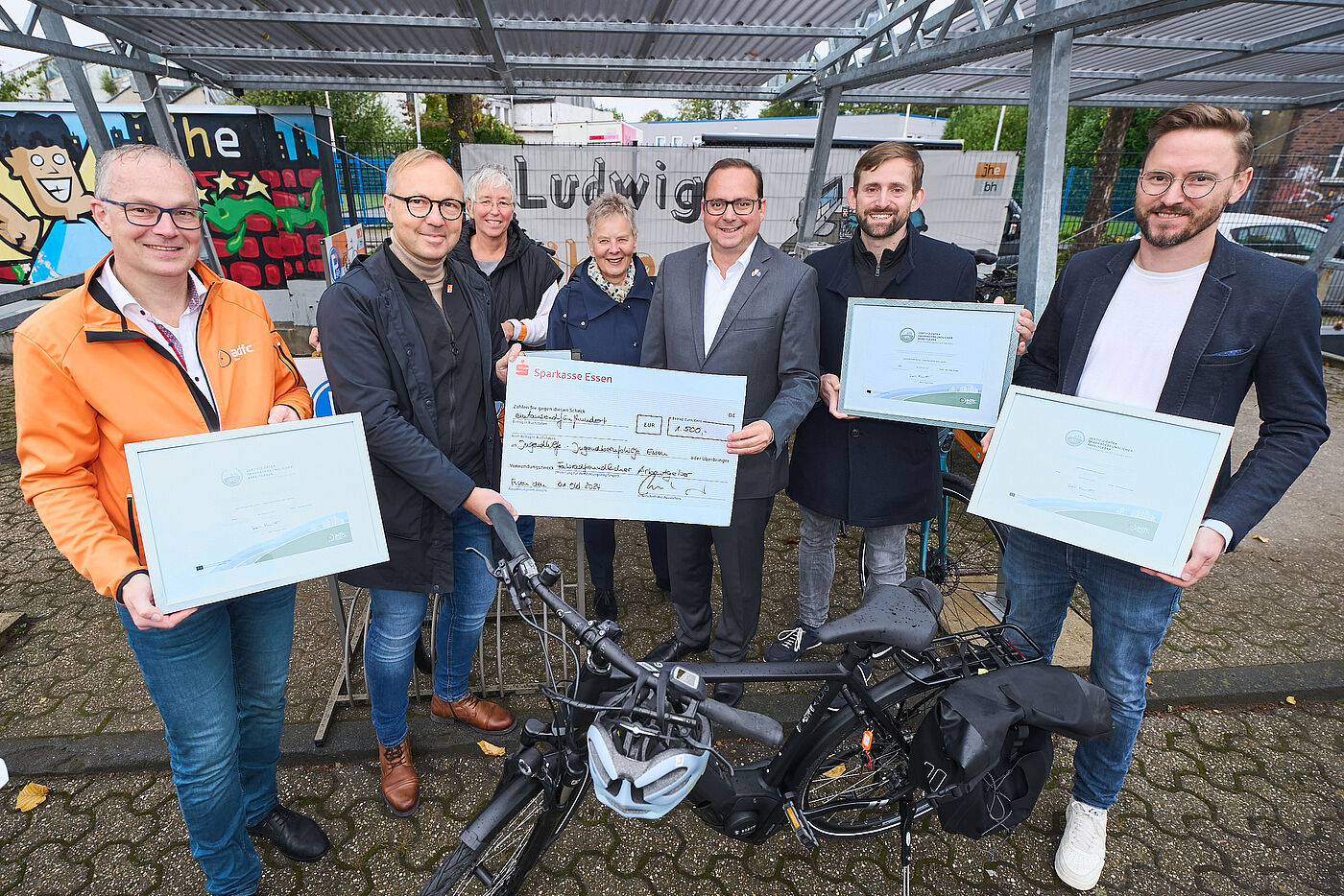 Auf dem Bild sind sieben Personen zu sehen. Einige von Ihnen halten gerahmte Urkunden in die Kamera. Im Vordergrund des Bildes steht ein Fahrrad.