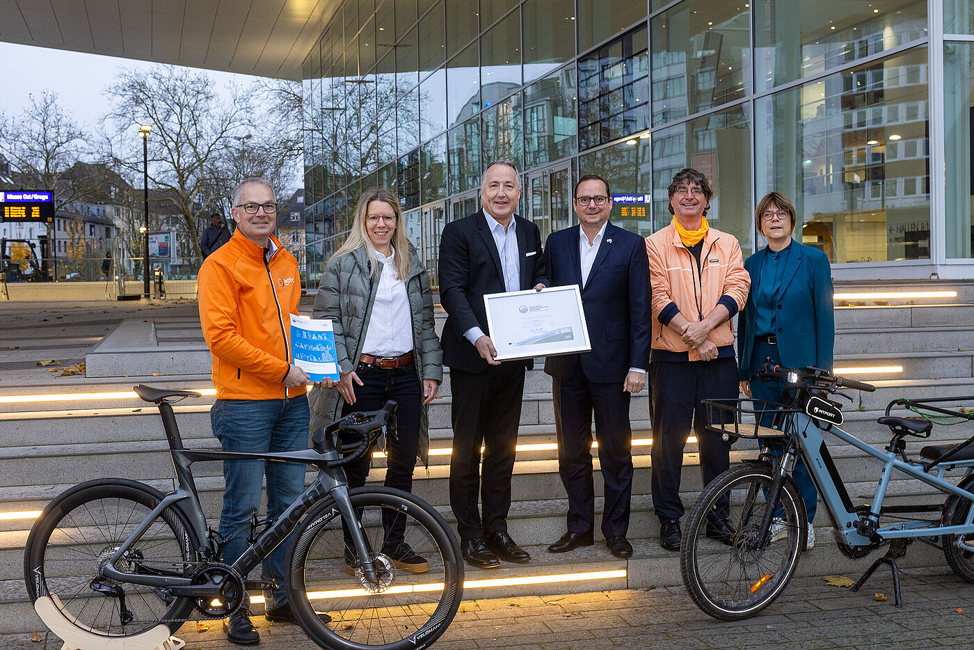 Sechs Personen stehen vor einem Gebäude mit Glasfassade. In der Mitte stehen zwei Männer, die gemeinsam ein gerahmtes Zertifikat hochhalten. Im Vordergrund stehen zwei Fahrräder.