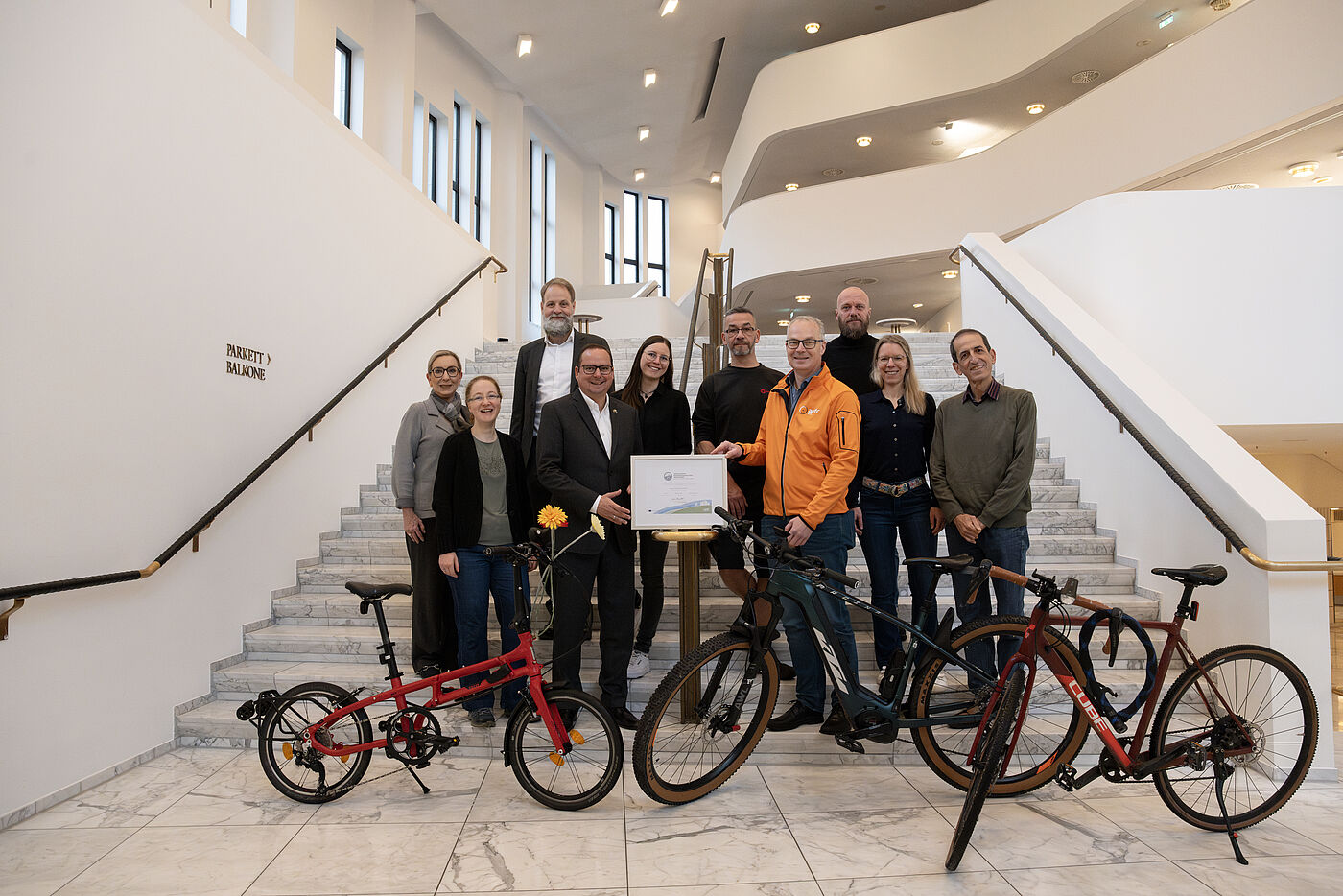 Zehn Menschen stehen auf einer weißen Treppe. Ein Mann in Anzug und ein Mann in einer orangenen Jacke halten ein gerahmtes Zertifikat. Die anderen Menschen stehen um die beiden Männer herum. Im Vordergrund sind Fahrräder zu sehen. Im Hintergrund sieht man weitere Treppen und Ballustraden.