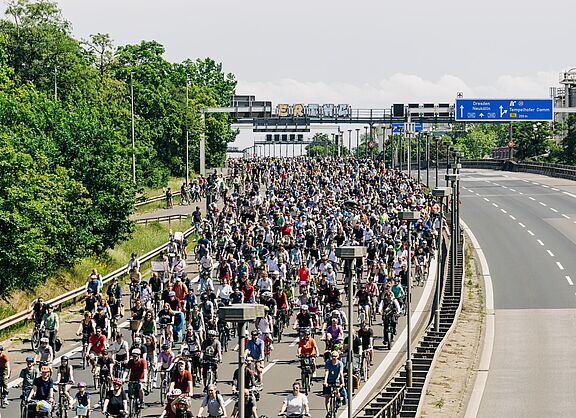 Eine große Menschenmenge fährt auf Fahrrädern über eine mehrspurige Autobahn, die bis zum Horizont mit Radfahrenden gefüllt ist. Über der Straße befinden sich Verkehrsschilder, die unter anderem Richtung „Dresden“ und „Neukölln“ zeigen. Links säumen Bäume die Straße, rechts ist eine leere Gegenfahrbahn zu sehen.